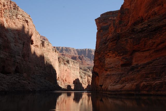 A scenic view of a narrow canyon with steep reddish rock formations, reflecting in a calm river under a clear blue sky.